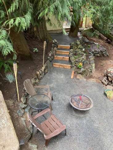 This image shows a cozy outdoor seating area with two chairs, a small glass table, a fire pit, stone steps, tiki torches, and large trees surrounding the space.