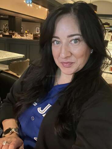 A woman with long dark hair smiles for a selfie in a restaurant. She wears a blue shirt, black blazer, and gold watch. The setting is elegant.
