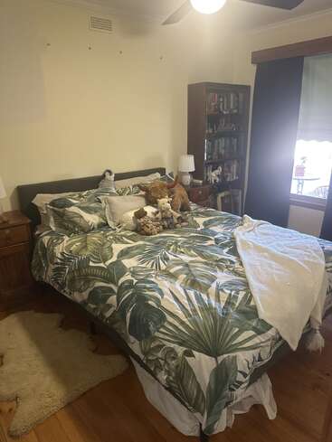 A cozy bedroom with a leafy-patterned bedspread, several plush toys on the bed, wooden furniture, bookshelf, a sheepskin rug, and natural light from the window.