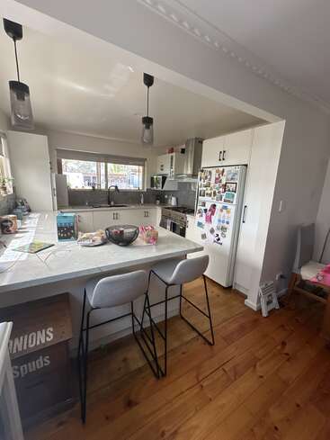 A modern kitchen features white cabinets, a countertop with snacks, two stools, hanging lights, a refrigerator covered in magnets, and wooden floors, creating a cozy atmosphere.