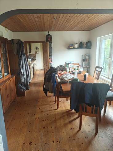 Ein gemütliches Esszimmer mit einem Holzboden und einer Holzdecke, einem vollgestellten Tisch, Mänteln auf den Stühlen, natürlichem Licht durch große Fenster und einem Blick in die Küche.