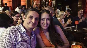 A smiling couple sits closely together in a lively, crowded restaurant with warm lighting. The atmosphere appears cheerful, social, and filled with conversations and laughter around them.