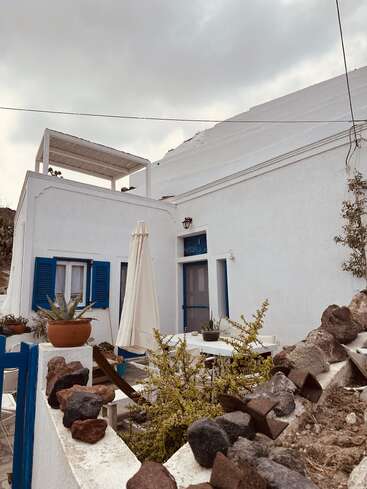 The image depicts a white building with blue accents, featuring a patio area with a table, umbrella, and potted plants, set against a cloudy sky backdrop.