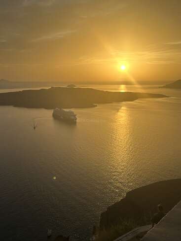 A peaceful sunset over calm waters, casting a golden hue. A cruise ship sails near dark islands, while a lone person admires the breathtaking view.