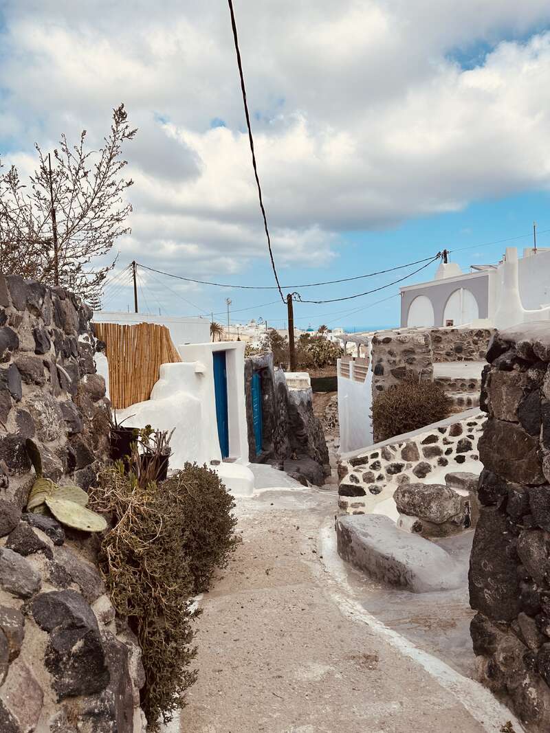 The image depicts a narrow, winding path in a small village, flanked by stone walls and featuring white buildings, with a blue door and a cloudy sky in the background.