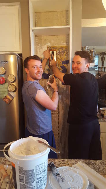 Two men are tiling a kitchen wall, standing side by side, with a bucket of white grout on the counter in front of them.