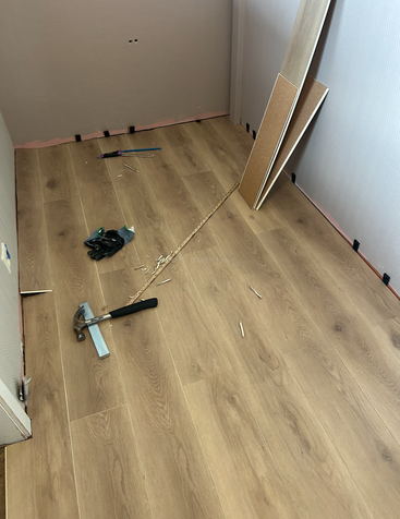 A small room is undergoing laminate flooring installation. Tools, gloves, and floorboards are scattered across the unfinished wooden floor, indicating ongoing renovation work and effort.