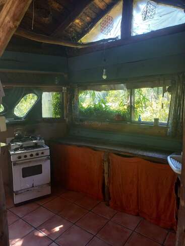 Esta acogedora cocina rústica cuenta con una estufa vintage, vigas de madera natural, suelo de baldosas, plantas en el alféizar y cortinas bajo la encimera, que crean un ambiente cálido y terrenal.