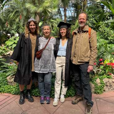 The image depicts four individuals standing together in front of a lush garden backdrop, with two wearing graduation attire, conveying a celebratory atmosphere.