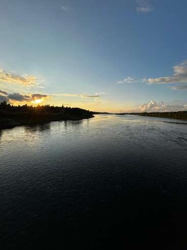Das Bild zeigt einen ruhigen Fluss, der unter einem klaren blauen Himmel bei Sonnenuntergang fließt, während das Sonnenlicht durch die Bäume scheint und sich auf dem ruhigen Wasser spiegelt. Eine friedliche Landschaft.