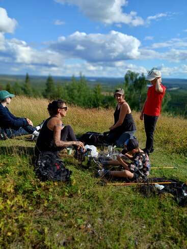 Eine Gruppe Leute sitzt entspannt auf einem grasbewachsenen Hügel und genießt gemeinsam ein Picknick unter einem wunderschönen blauen Himmel mit flauschigen Wolken und einer malerischen Aussicht.