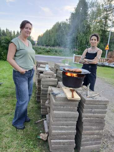 Zwei Frauen stehen neben einem provisorischen Grill aus aufgestapelten Ziegelsteinen und braten Würstchen über offenem Feuer, umgeben von viel Grün und einer ruhigen Waldlandschaft.