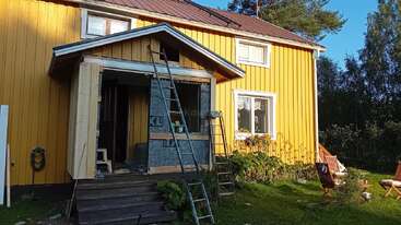 Auf dem Bild sieht man ein gelbes Holzhaus, an dem gerade eine neue Veranda gebaut wird. An dem Haus lehnen Leitern. Im Garten stehen Stühle und es gibt viel Grün.