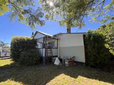 A small weatherboard house is surrounded by green bushes and trees. A ladder leans on the porch. The sun shines brightly through leafy branches above.