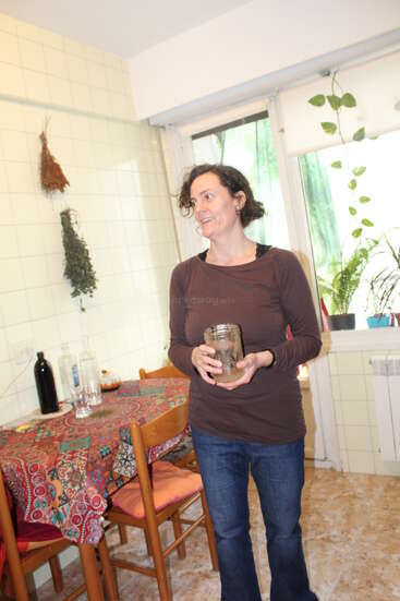 Uma mulher está em pé numa cozinha bem iluminada, segurando um pote e sorrindo. Atrás dela, há uma toalha de mesa colorida, cadeiras, ervas penduradas e vários vasos de plantas perto da janela.