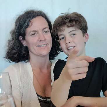 A woman with curly hair smiles while sitting next to a young boy. The boy is pointing towards the camera. Both appear happy and relaxed together.