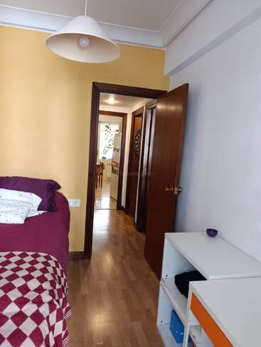A imagem mostra um pequeno quarto com uma cama marrom-avermelhada, estantes brancas, piso de madeira, paredes amarelas e brancas e uma porta aberta que dá para um corredor.
