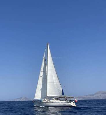 Un velero blanco se desliza por un mar azul profundo bajo un cielo brillante y despejado. Montañas lejanas se desvanecen en el horizonte, evocando un viaje tranquilo y lleno de aventuras.