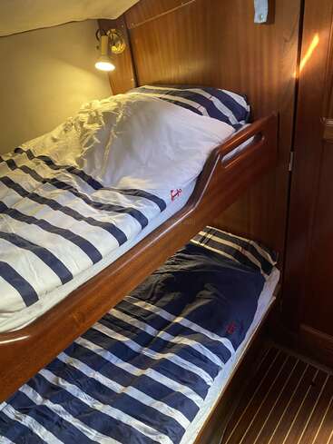 Esta imagen muestra una acogedora cabaña de madera con dos literas, ropa de cama a rayas náuticas, una almohada, una lámpara de lectura y paredes de madera, que dan un ambiente cálido y acogedor.