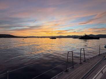 Una preciosa puesta de sol sobre un mar en calma, vista desde un barco. Varios veleros y yates descansan sobre el agua, mientras unas nubes de colores iluminan el cielo.