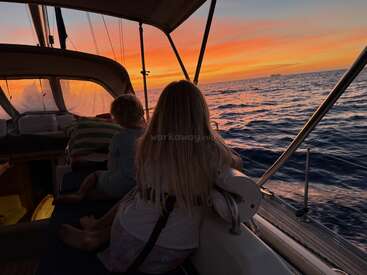 Dos niños están sentados en un velero al atardecer, contemplando el cielo teñido de tonos anaranjados y morados sobre el mar en calma, disfrutando de un momento de tranquilidad.