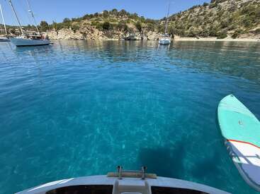 Unas aguas turquesas y cristalinas rodean una pequeña embarcación y una tabla de paddle surf, con veleros fondeados cerca. Colinas rocosas cubiertas de árboles verdes se alzan bajo un cielo azul brillante. Una escena tranquila.