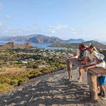 Una familia de cuatro miembros disfruta de una vista panorámica desde la ladera de una montaña, con vistas a una ciudad costera de verdes colinas, mar azul e islas lejanas bajo un cielo soleado.