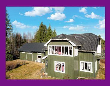 Dieses Bild zeigt ein grünes Holzhaus und ein Nebengebäude unter einem strahlend blauen Himmel mit vereinzelten Wolken, umgeben von Bäumen und Gras, eingerahmt von einem lila Rand.