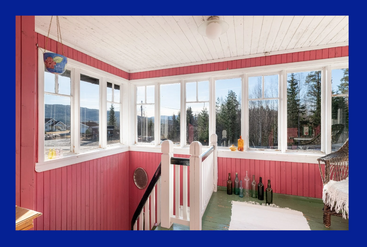 Dieser sonnendurchflutete Raum hat rosafarbene Holzwände, viele große Fenster mit Blick nach draußen, eine weiße Decke, einen grünen Boden, Glasflaschen, einen geflochtenen Stuhl, eine Hängepflanze und einen Teppich.