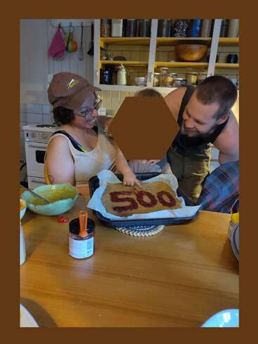Eine fröhliche Familie feiert zusammen in einer gemütlichen Küche und lächelt über einen selbstgebackenen Kuchen, der mit der roten Zahl "500" verziert ist und einen besonderen Meilenstein oder eine besondere Leistung hervorhebt.