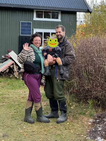 A smiling couple stands outside, one waving. The man holds a child with a frog emoji covering their face. All wear boots near a green house.