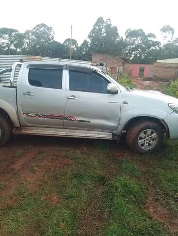 Un pick-up argenté portant des autocollants "TRD Sportivo" est garé sur un terrain herbeux et boueux, à proximité de bâtiments ruraux, entouré d'arbres et d'un ciel couvert.