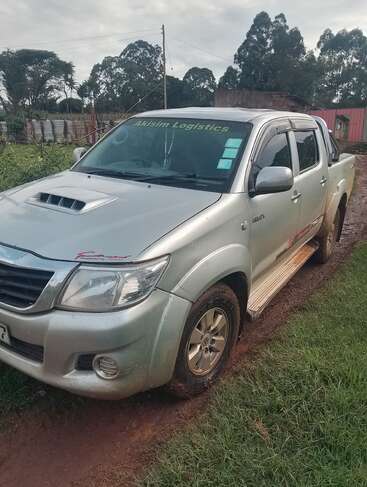 Une camionnette Toyota Hilux argentée, portant l'inscription "Akisim Logistics" sur le pare-brise, est garée sur un chemin boueux, entouré de verdure et de clôtures rurales.