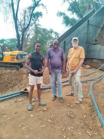 Trois hommes se tiennent ensemble sur un chantier de construction ou d'exploitation minière avec des tuyaux, de la terre et des machines. Des arbres et des équipements sont visibles à l'arrière-plan. Tenue décontractée, visages souriants.