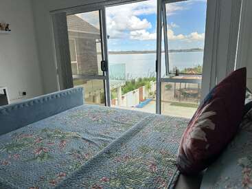 A cozy bedroom with a floral quilted bedspread overlooks a serene lake through large glass windows, featuring outdoor seating and clear skies with fluffy clouds.
