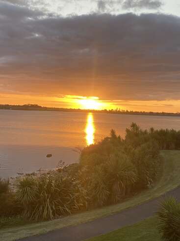 A golden sunset reflects beautifully on calm water, framed by lush green plants, a winding path, and dramatic clouds hanging above the peaceful scene.