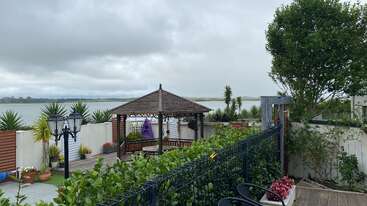A beautifully landscaped garden with lush greenery, outdoor gazebo, potted plants, black fence, and view of a serene lake under a cloudy, overcast sky. Peaceful atmosphere.
