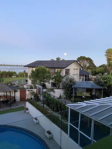 A serene backyard features a swimming pool, gazebo, lush green garden, and patio. A house overlooks a calm body of water under a clear evening sky.