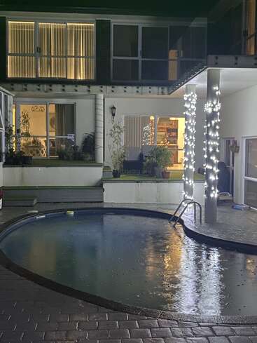 A cozy backyard at night, featuring a small, illuminated swimming pool, patio lights wrapped around columns, potted plants, and a warmly lit house interior.
