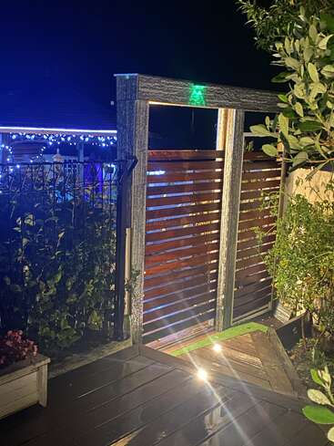 A wooden gate framed in metal opens onto a lit, wet deck at night. Festive blue and white lights decorate a gazebo in the background. Lush greenery surrounds.