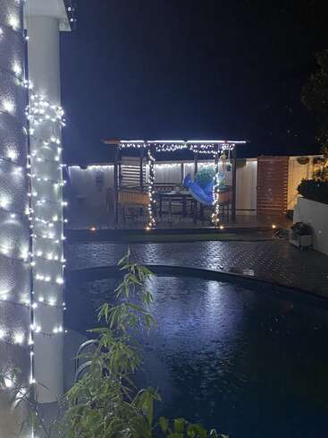 A cozy backyard scene at night, featuring a gazebo decorated with string lights, overlooking a pool. Plants and festive lights create a warm, inviting atmosphere.