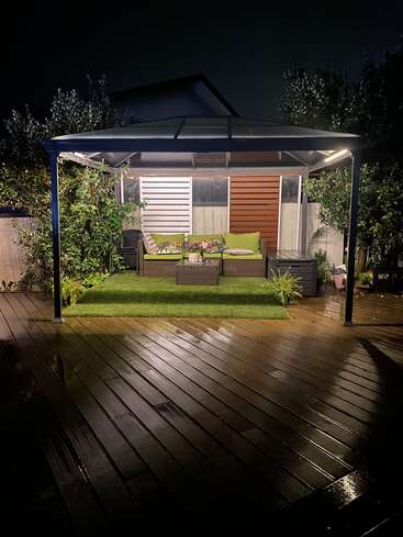 A cozy outdoor seating area under a gazebo, illuminated at night, with green artificial grass, colorful cushions, and wet wooden decking reflecting the soft light.