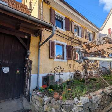 Uma casa amarela charmosa com persianas marrons, detalhes rústicos em madeira, um pequeno canteiro de pedra, flores da primavera, trepadeiras e decorações caprichosas de jardim sob um céu azul.