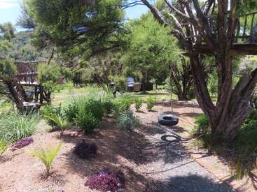 A peaceful garden scene with lush greenery, a wooden treehouse, a hanging tire swing, scattered shrubs, and sunlight filtering through the trees, creating a serene atmosphere.