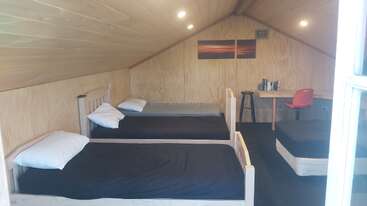 This cozy wooden room features two single beds with black bedding, a small desk with a red chair, a stool, books, and warm overhead lighting.