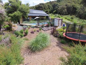 This image shows a countryside backyard with a swimming pool, fenced area, garden plants, a trampoline, a shed, and lush green trees surrounding the property.