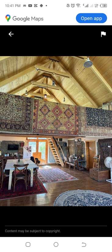 This cozy room features a high wooden ceiling, vibrant patterned rugs on walls and floors, wooden furniture, a staircase, warm lighting, and eclectic decorations throughout.