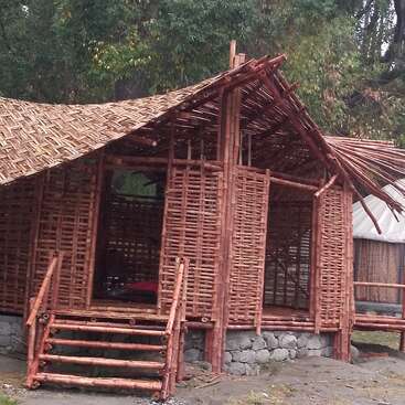 This image shows a rustic bamboo hut with a woven bamboo roof and walls, elevated on a stone foundation, surrounded by greenery and trees outdoors.