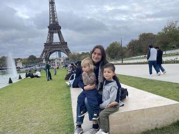 A woman and two children sit on a stone bench near the Eiffel Tower in Paris, enjoying a cloudy day with a fountain and people around.
