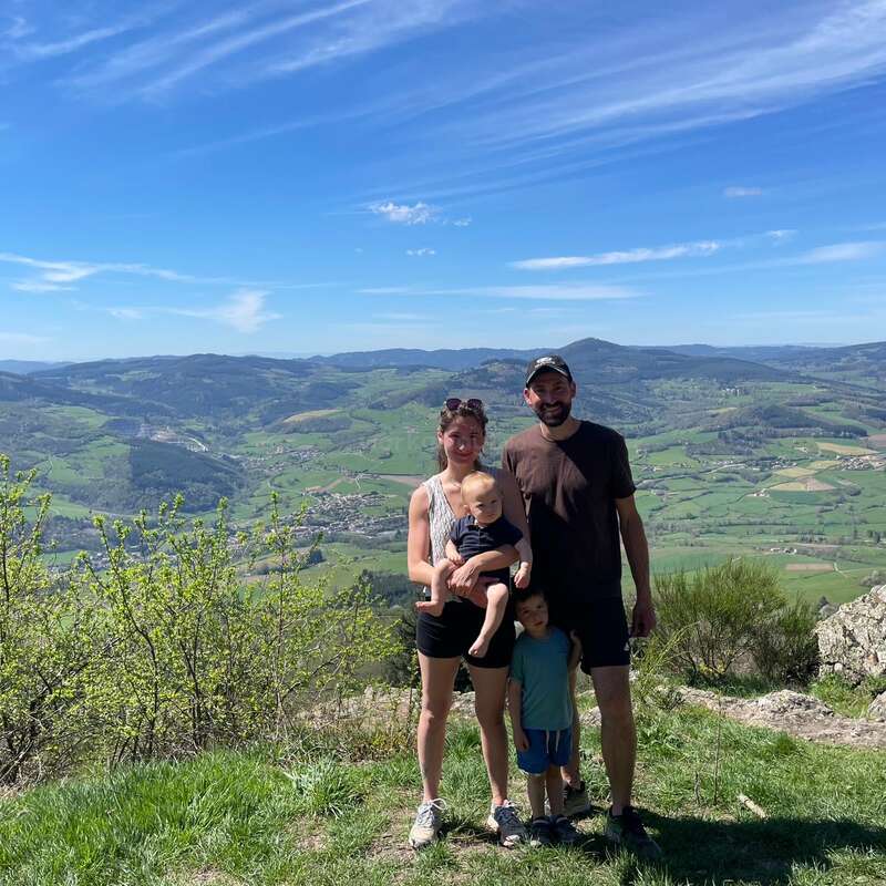 The image depicts a family of three standing on a hilltop, with a picturesque landscape of rolling hills and mountains in the background under a clear blue sky.